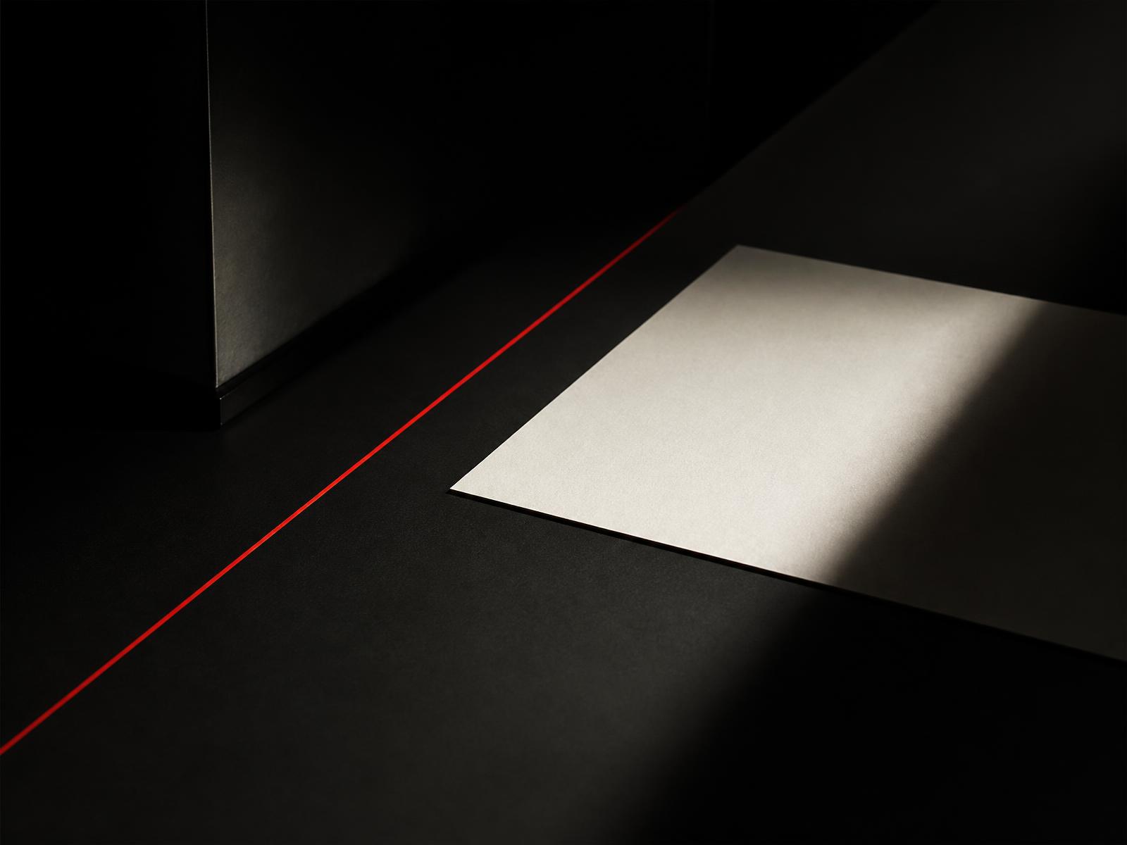 A single cream sheet of paper lit by hard directional light on a near-black desk, crossed by a thin red line